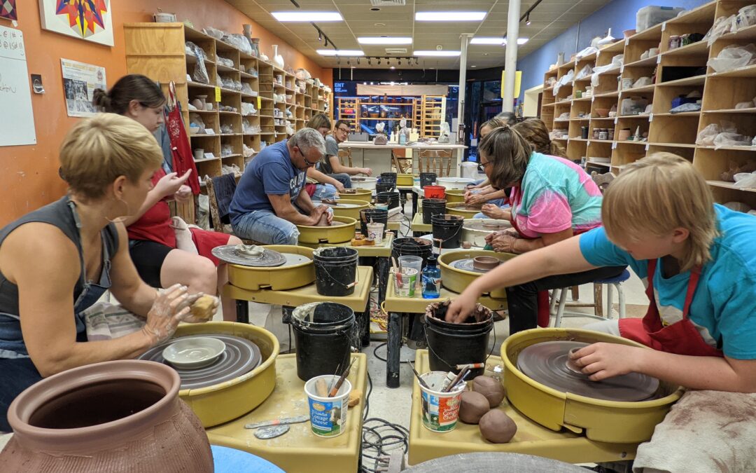 Class of 12 students all ages observing their instructor demonstrate throwing on the pottery wheel