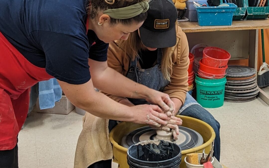 Instructor helping student learn to throw on the potter's wheel by placing her hands over their hands to show proper hand placement.